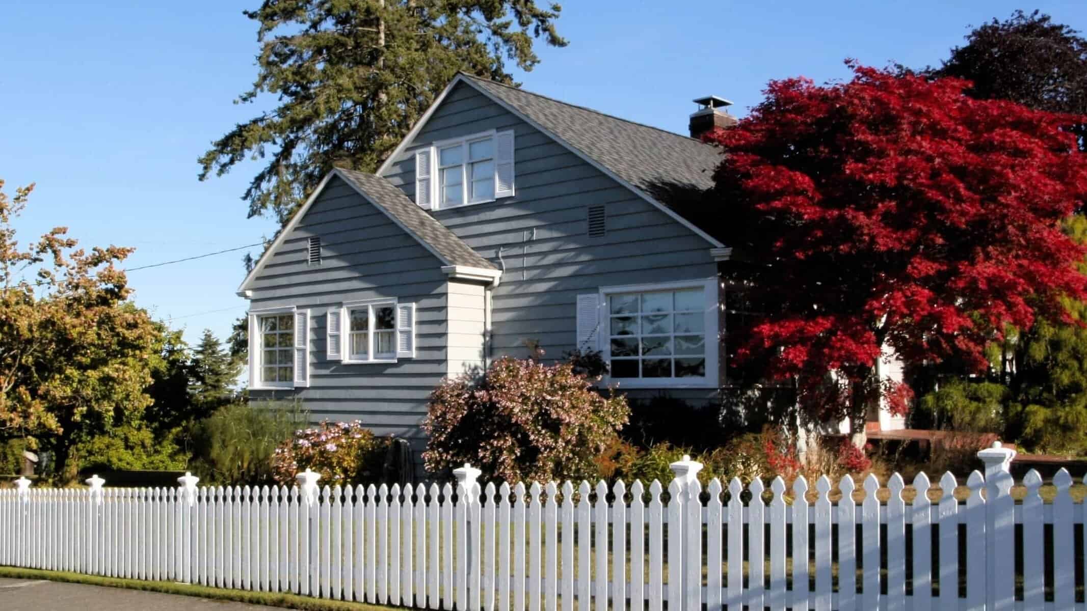 A two-story gray house with white trim, surrounded by lush greenery and a vibrant red tree, stands behind a white picket fence under a clear blue sky.