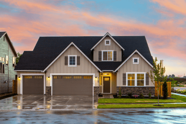 A modern two-story house with a gabled roof, three-car garage, and lit entrance under a colorful sunset sky.