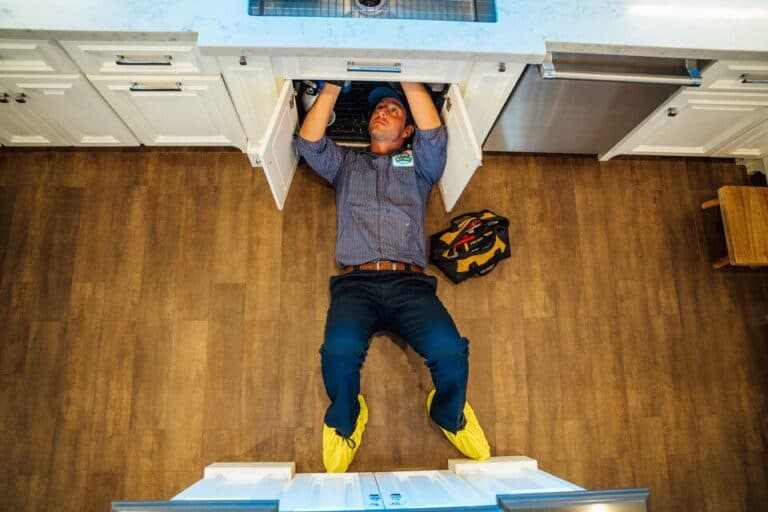 A worker lies on the floor under a kitchen sink, wearing yellow shoe covers and working with tools on a plumbing repair, while a tool bag sits nearby.