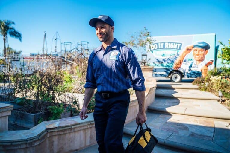 A man in a uniform carrying a tool bag walks outdoors near a “Do It Right” truck on a sunny day, ready for plumbing repair.