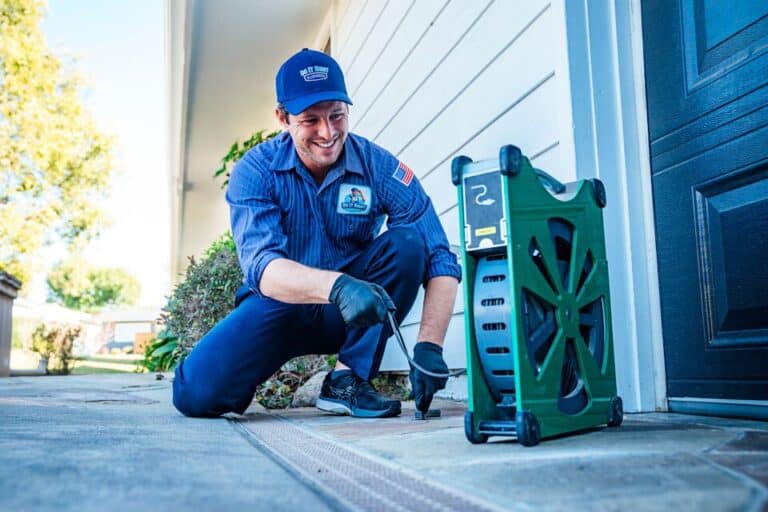 A technician in uniform kneels by a house, using a tool near the ground with a green equipment box beside him, expertly handling water line installation tasks.
