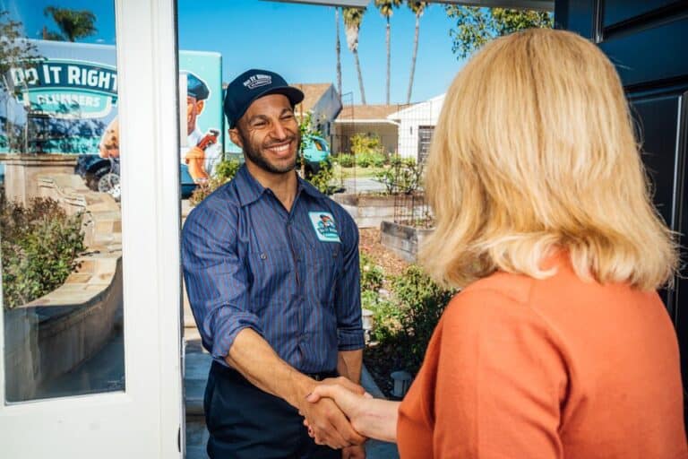 A smiling uniformed worker shakes hands with a woman at the door of a house on a sunny day, ready to discuss water line installation.