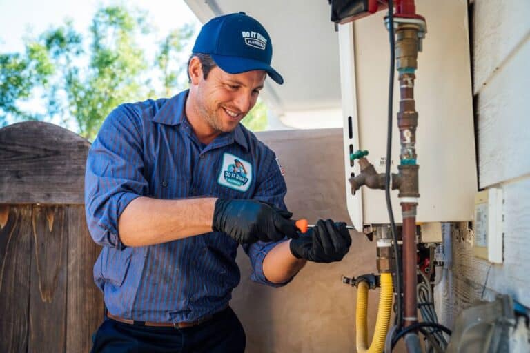 A plumber wearing gloves and a uniform uses a screwdriver to work on outdoor plumbing equipment, focusing on water line installation attached to a house.