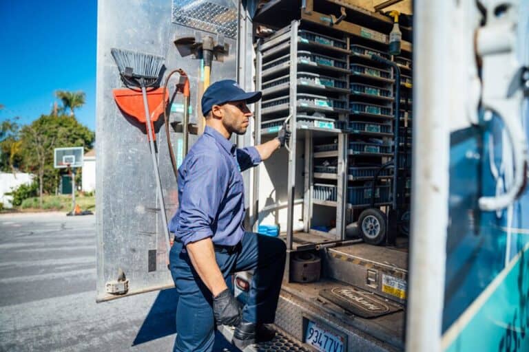 A man in work attire loads or unloads trays labeled Tankless Water Heater Maintenance from a delivery truck parked on a street in a residential area.