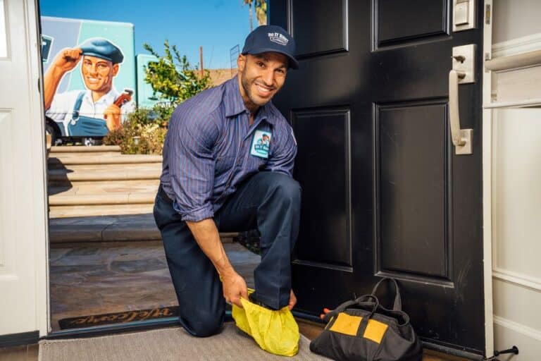 A smiling technician kneels at a doorway, putting on yellow shoe covers for a Tankless Water Heater Maintenance visit, with a tool bag nearby and the company truck visible outside.