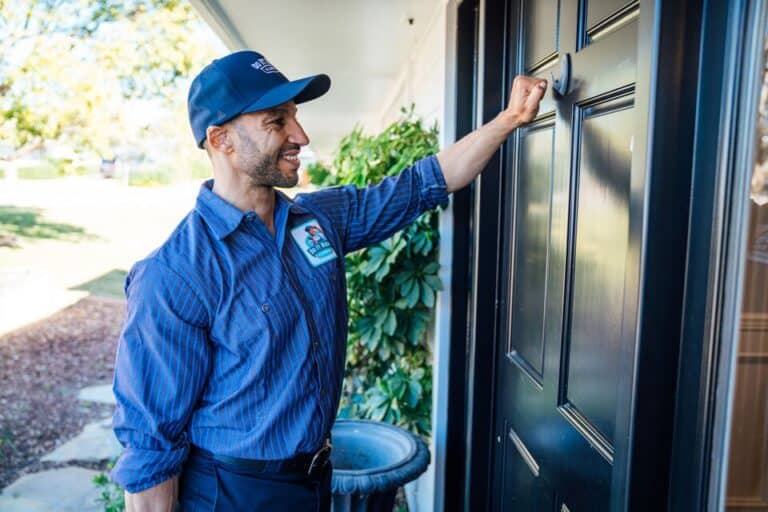A delivery worker in uniform, specializing in tankless water heater maintenance, knocks on a black front door with a smile, greenery visible in the background.