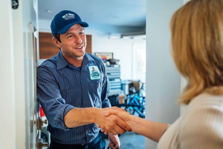 A repair technician in uniform, specializing in Tankless Water Heater Maintenance, shakes hands with a woman at her front door, standing in a garage or entryway.