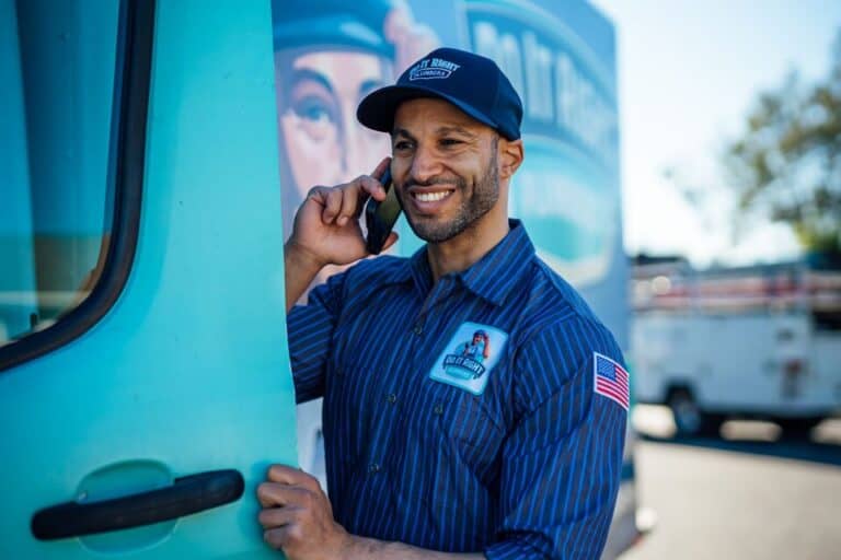 A man in a blue uniform and cap stands by a van, smiling while talking on a smartphone. An American flag patch is visible on his sleeve, suggesting he might be discussing tankless water heater maintenance services with a customer.