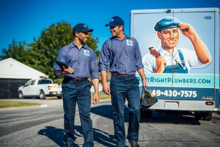 Two plumbers in uniform walk beside a company van featuring a large illustration of a plumber holding a wrench, the business name, contact information, and an ad for expert Tankless Water Heater Maintenance.