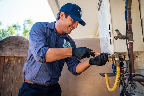 A technician wearing gloves and a uniform uses a screwdriver to work on an outdoor tankless water heater mounted on a wall.