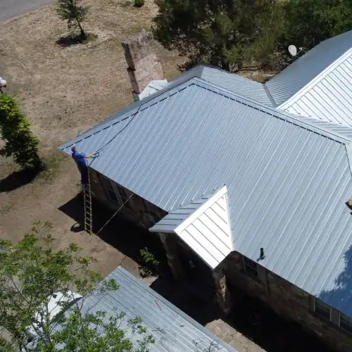 Technician on ladder installing metal roof panels on stone house, illustrating cost of metal roof San Antonio by Dolan Roofing