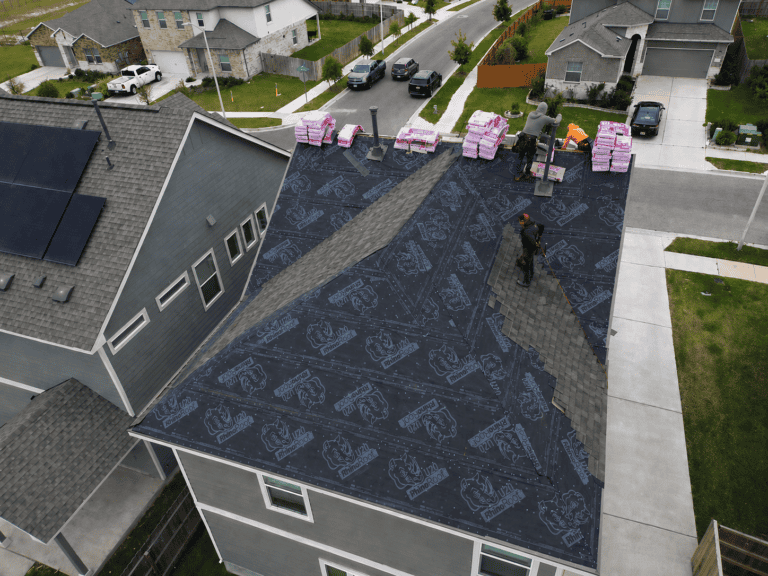 Residential house roof under construction with black RhinoRoof underlayment partially installed; workers placing asphalt shingles while bundles of shingles and tools are visible near the ridge, with neighboring homes in the background in San Antonio