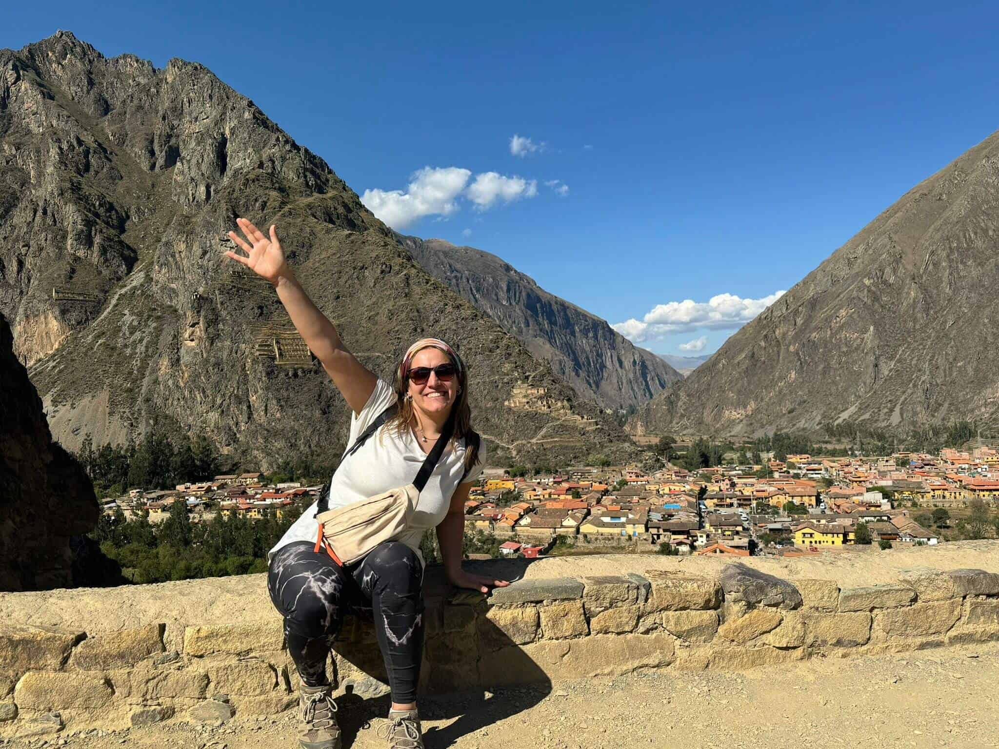 Vibrant woman smiling and waving in Machu Picchu with mountains and ancient ruins in the background.