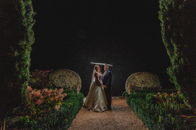 Deer Park Devon Wedding -Stunning bride and groom under umbrella in rain at night, surrounded by illuminated garden, captured in a romantic wedding photo by Eyecandy Weddings - Photo + Video.
