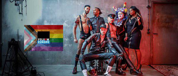 Gruppe von Menschen beim Folsom Berlin 2025, Pride-Event mit bunten Outfits und LGBTQ+-Flagge im Hintergrund.