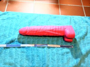 Das Bild zeigt einen roten Gleitstab aus Silikon, der auf einem blauen Handtuch liegt. Ein Messlineal befindet sich darunter, um die L&auml;nge des Gleitstabs zu verdeutlichen. Das Bild ist Teil eines Beitrags auf der Website Fistwerk, der sich mit Produkten und Zubeh&ouml;r f&uuml;r Fisting besch&auml;ftigt.