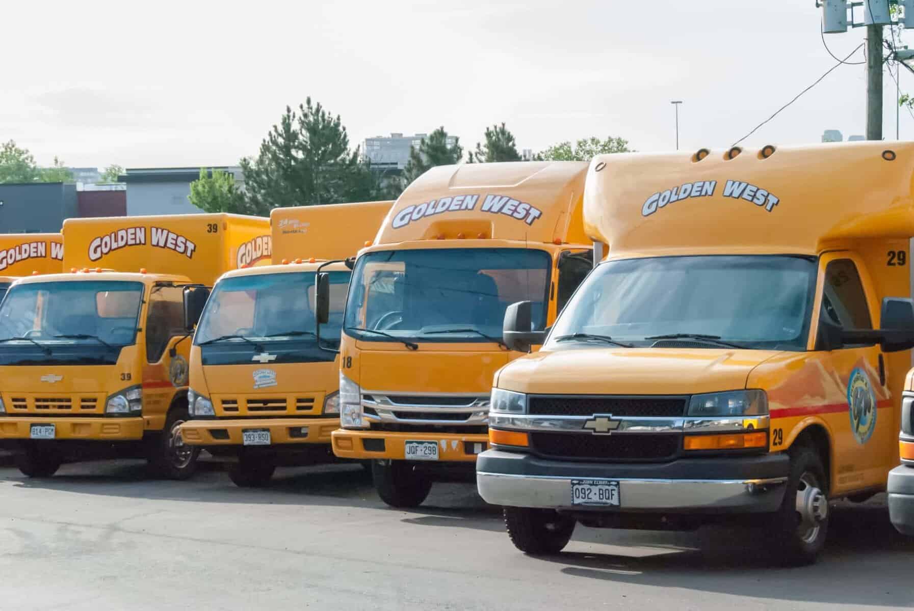 Five yellow Golden West shuttle buses are parked in a row within their service area lot, with trees and a building visible in the background.
