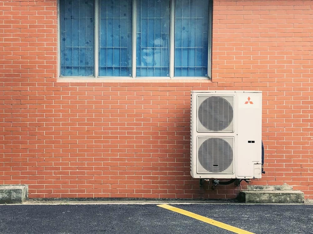 A white air conditioning unit is mounted on a red brick wall beneath a blue-barred window, next to a black asphalt parking lot with a yellow line—a frequent sight for Denver homeowners facing AC problems.