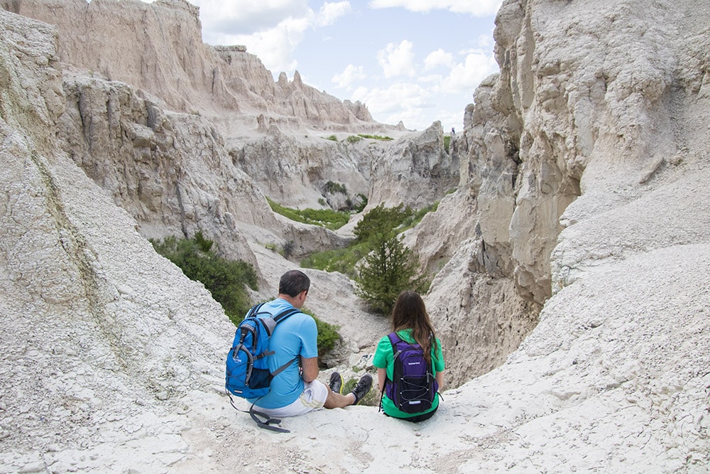 Discover the Funnest National Parks for Kids Dad and daughter enjoying the panoramic view of the Badlands from a hiking trail. They sit together, admiring the unique rock formations and rugged terrain. The image captures the bond between them and the vast beauty of Badlands National Park.