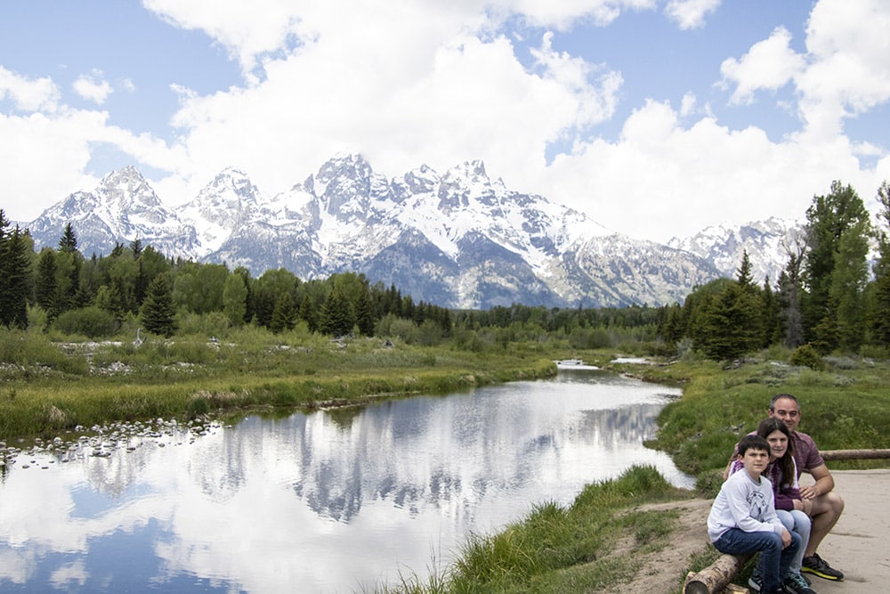The Best Funny Hiking Quotes For Instagram (Captions + Puns Too) Family hiking in Grand Teton National Park sitting on a log in front of a river that shows the reflection of the snow-capped mountains.
