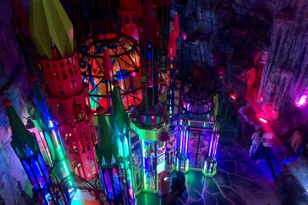 Meow Wolf Denver: The Ultimate Experience Guide for All Ages A towering castle structure with turrets and colorful lighting stands in the center of a large room. The castle is adorned with intricate details, including arches and windows with stained-glass accents. A small bridge extends from the castle, leading to a pathway on the opposite side of the room.