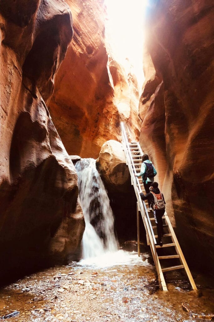 Kanarra Falls: Hike Utah’s Best Slot Canyon Two people climbing up the metal staircase at the first waterfall inside of the Kanarra Falls slot canyon. The ladder sits right next to the waterfall.