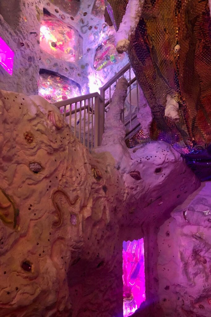 Meow Wolf Denver: The Ultimate Experience Guide for All Ages Altar-like structure in a dimly lit room with pink walls and floors at Meow Wolf Denver. The centerpiece of the room is a round table with candles, vases, and sculptures, surrounded by plush pink chairs. Various art installations and decor elements can be seen on the walls, including a large painting, a framed photo, and hanging pink tassels. The overall vibe of the room is mysterious and ethereal.