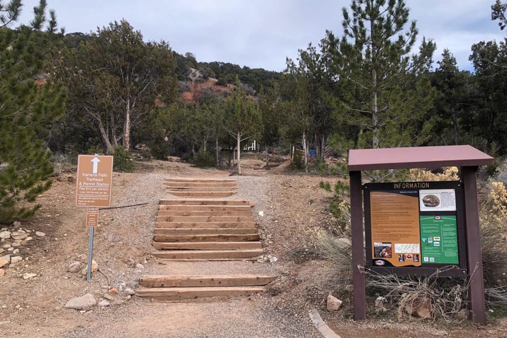 Kanarra Falls: Hike Utah’s Best Slot Canyon A view of the wooden stairs leading to the Kanarra Falls trailhead, showing a sturdy staircase built into a rocky hillside. The stairs have some landings leading up to a dirt path. The surrounding landscape features large evergreen trees.