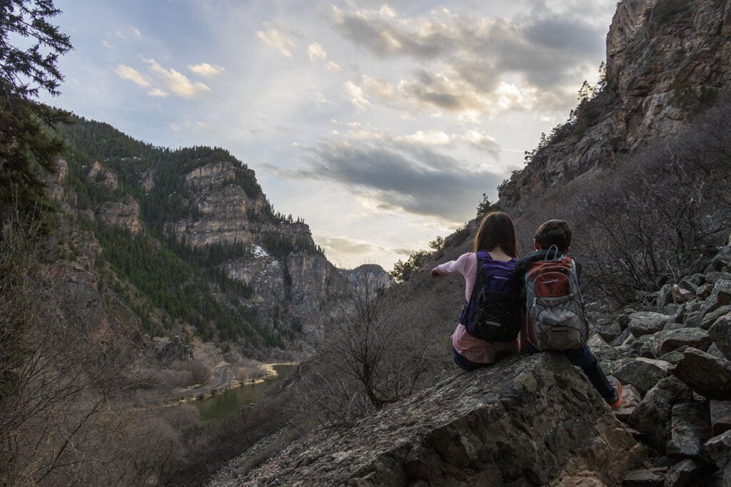 Hiking Hanging Lake in Colorado – The Ultimate Guide Kids sitting on a large boulder on the Hanging Lake trail overlooking the colorado river.