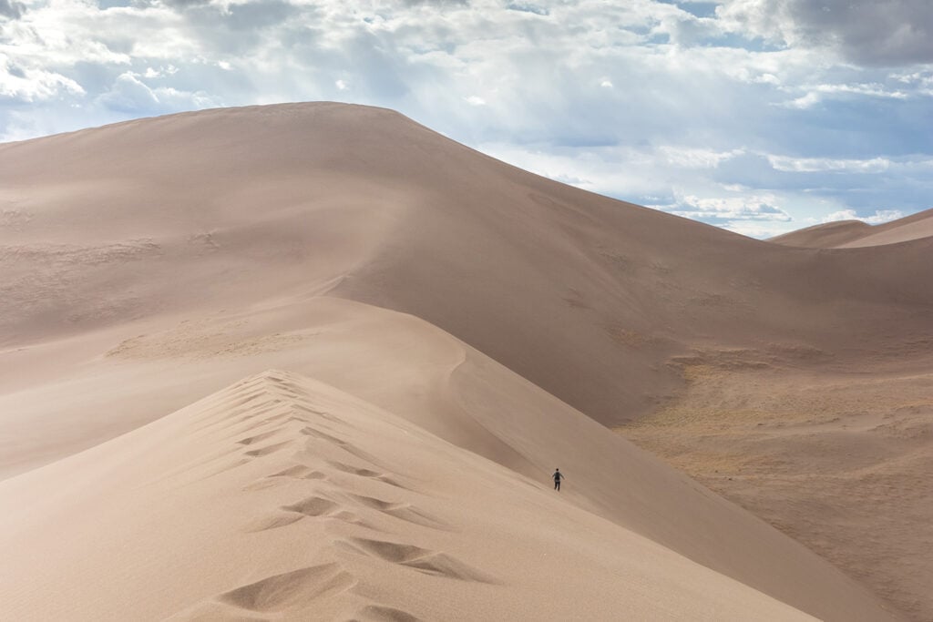 Discover the Funnest National Parks for Kids Great Sand Dunes National Park: Person walking on vast sand dunes, with blue skies in the background. A captivating desert landscape adventure.