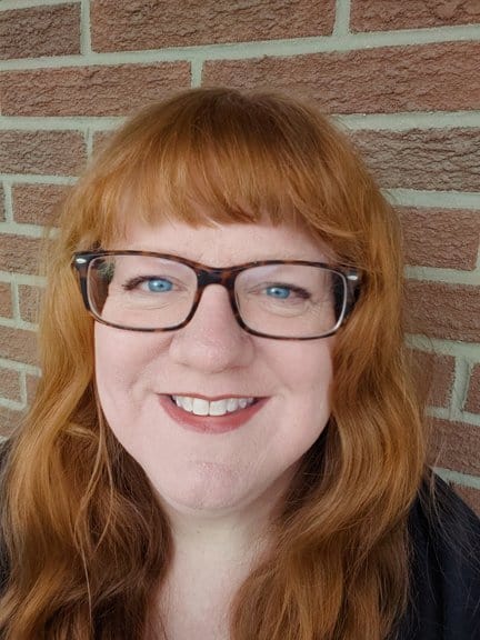 Supporter of Hero Initiative with red hair and glasses smiling against brick wall.