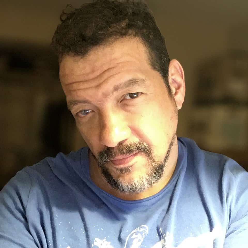 Close-up portrait of a middle-aged man with dark curly hair and a beard, wearing a blue shirt.