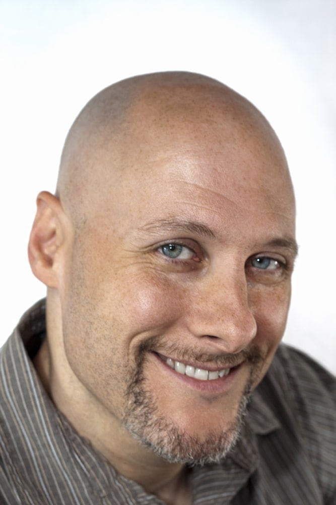 Close-up of a smiling, bald man with blue eyes and stubble, representing hope and resilience.