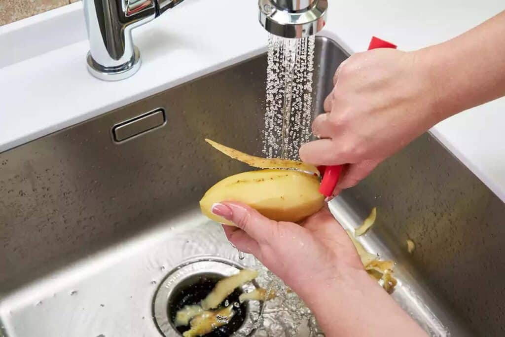 Person peeling a potato under running water in a kitchen sink.