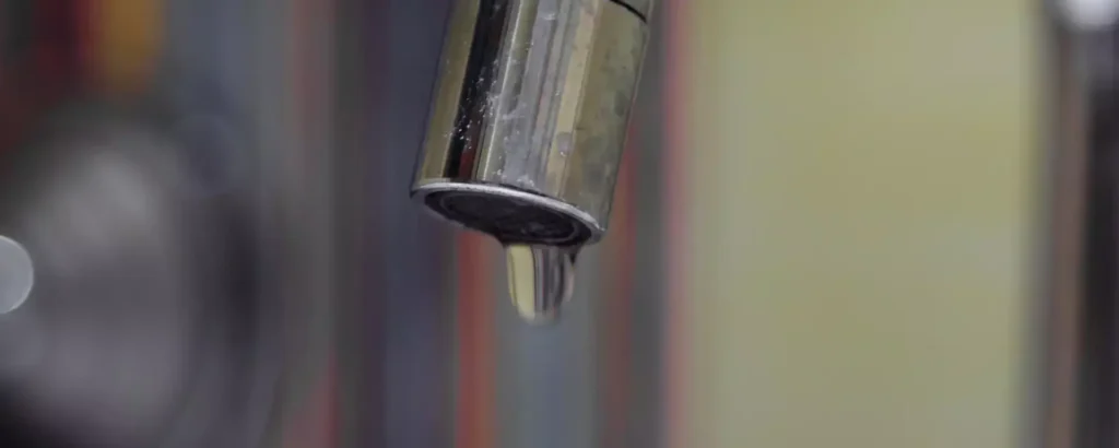A close-up of a kitchen faucet with a single water droplet hanging from the spout. The background is blurred with vertical stripes.