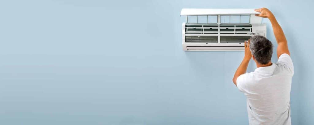 Person adjusting the front panel of a wall-mounted air conditioning unit against a light blue background.