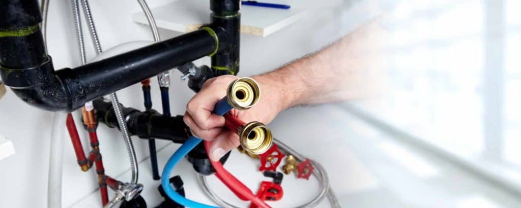 A person holding two plumbing hoses under a sink, surrounded by various pipes and fittings.