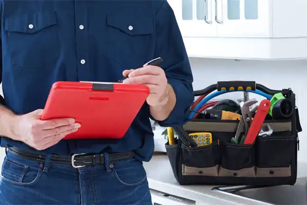 Person in a blue shirt holding a red clipboard stands near a kitchen counter with a tool bag full of various tools.