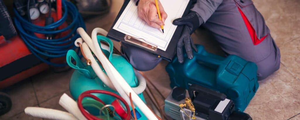 A technician in gray overalls kneels on the floor, writing on a clipboard. Nearby are tools, tubes, and a tank.