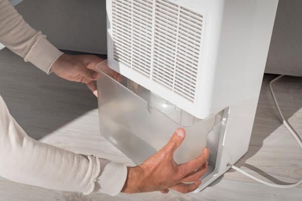 A person removing the water tank from a dehumidifier placed on a wooden floor.