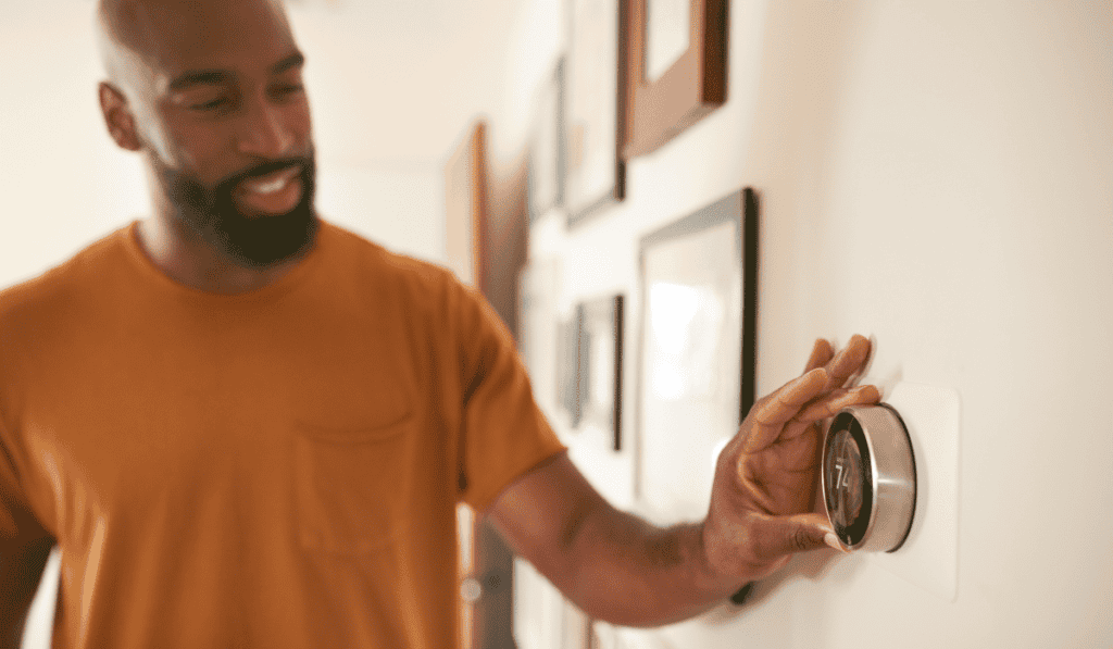 A man in an orange shirt smiles as he sets up a new smart thermostat on the wall, its display showing 74 degrees. Pictures hang nearby, capturing cozy moments.