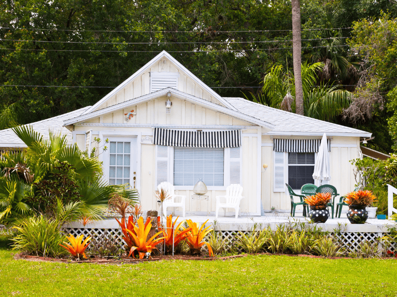 White cottage with striped awnings and a front porch that exudes charm. The cozy setup includes chairs and a table under an umbrella, perfect for enjoying vibrant orange plants in the garden. Lush trees provide shade, making it ideal for those seeking HVAC advice for older homes.