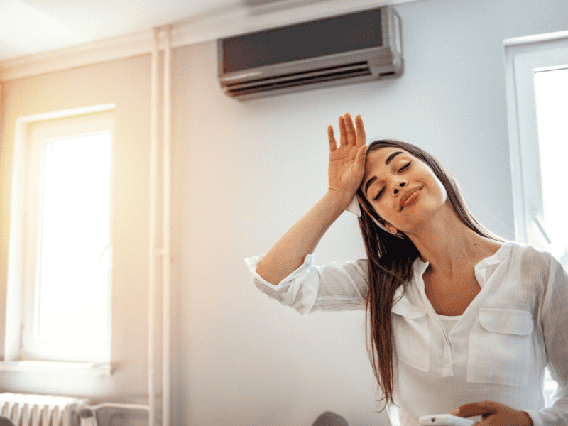 A woman in a white blouse stands in a room, holding her forehead with a relaxed expression. She enjoys the cool air from the AC unit beside her, hinting at one of her favorite tips for staying comfortable in older homes.