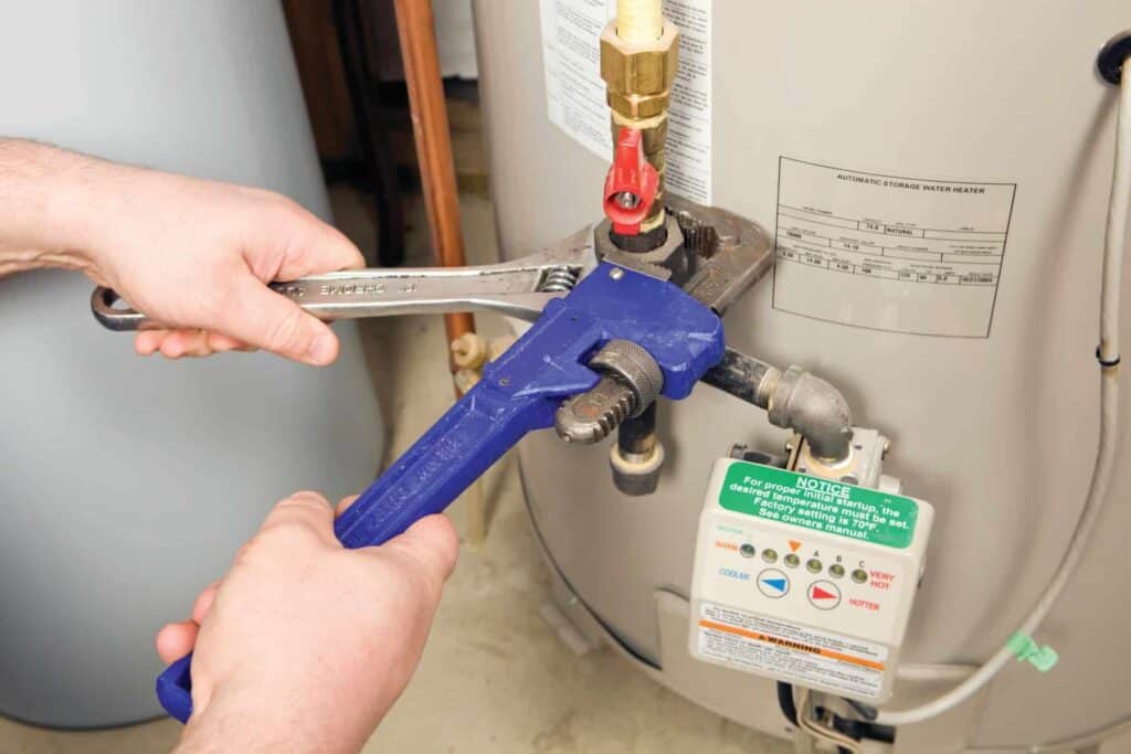 Two hands use wrenches to adjust a valve on a water heater. A notice hangs from the heater, providing instructions and color-coded guidelines.
