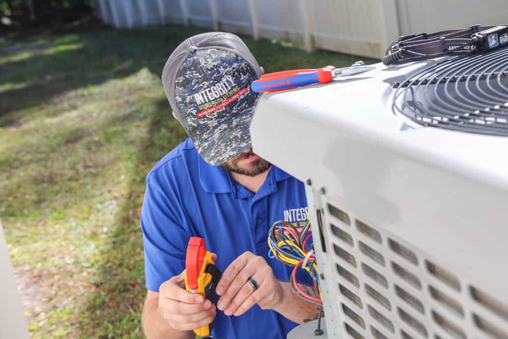 A technician in a blue shirt and cap uses tools to work on the wiring of an outdoor air conditioning unit.