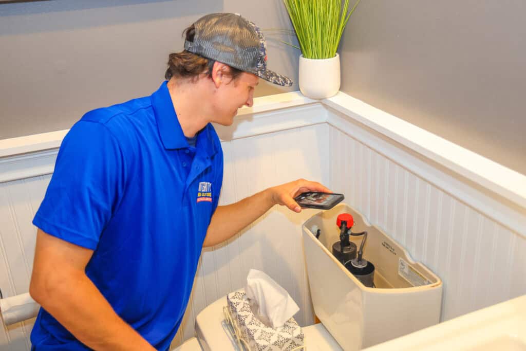 A man in a blue shirt examines the inside of a toilet tank, using his phone as a flashlight—a simple plumbing maintenance check that can help save money, especially during summer.