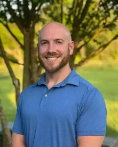 A Man With A Beard And Short Hair Smiles While Standing Outside In Front Of Trees, Wearing A Blue Polo Shirt.
