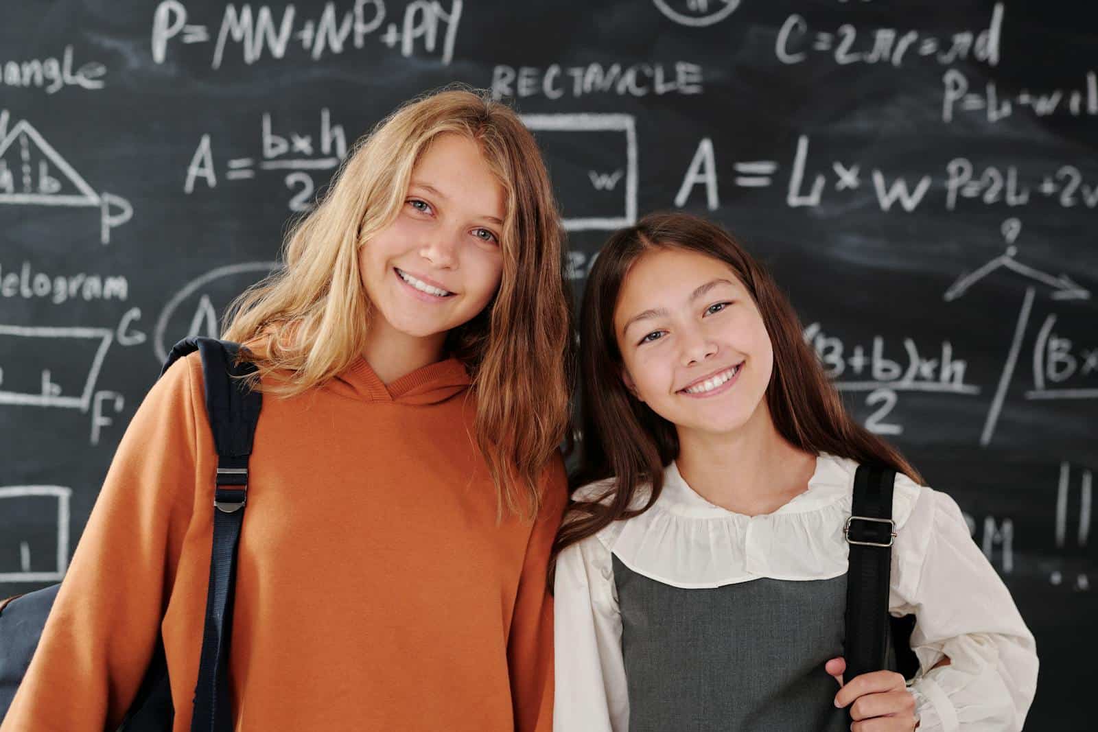 Photo by Max Fischer Two cheerful girls stand in a classroom with a math-covered chalkboard, ready for learning.