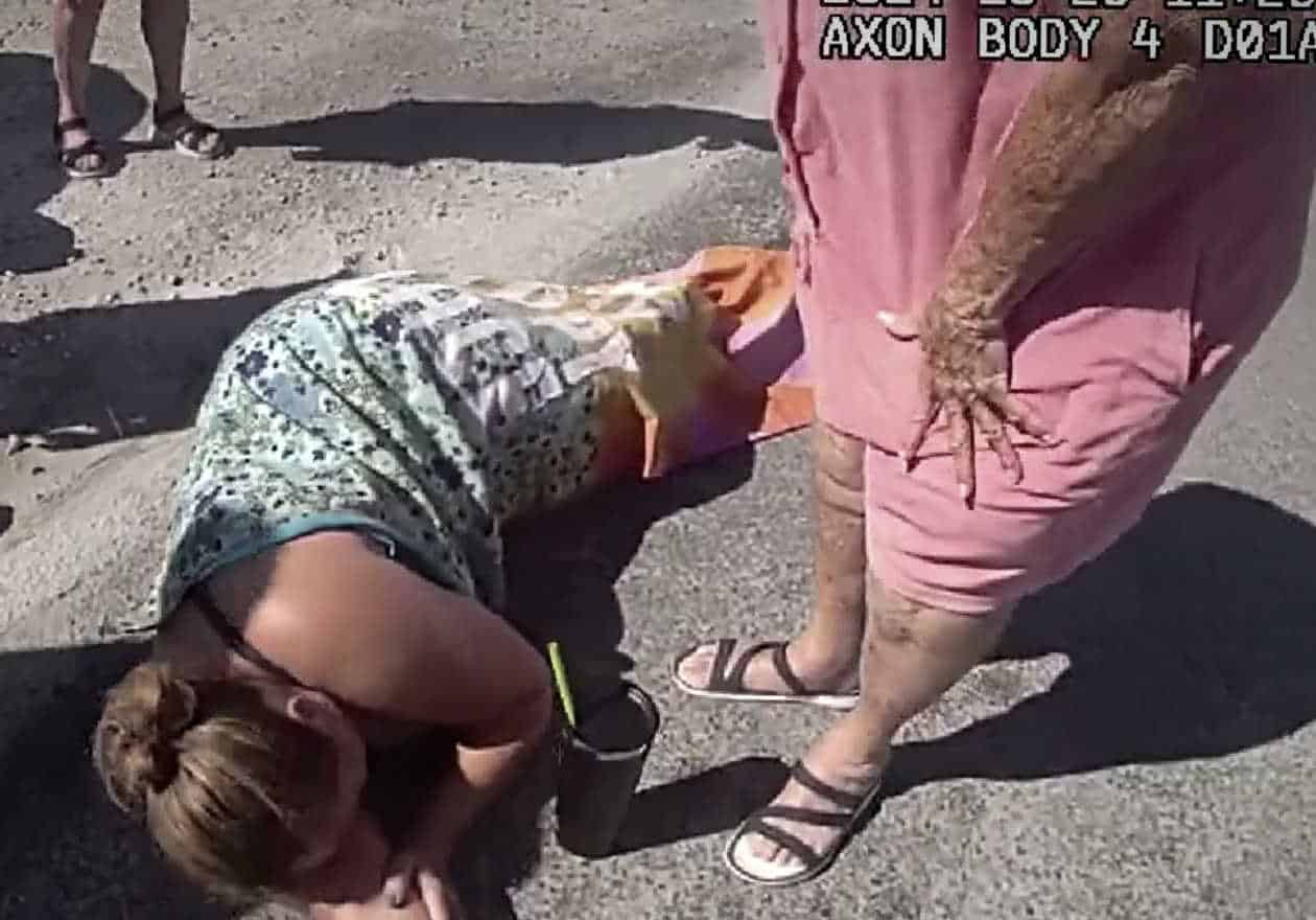 Injured woman lying on a marina pavement clutching her leg after being hit by a trailer, with concerned bystanders nearby. Image captured from police bodycam footage, showing timestamp overlay in the corner.