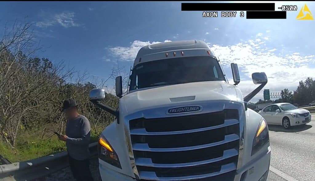 Police body-camera image showing the front of a white Freightliner semi-truck stopped on the highway shoulder, with a man standing near a guardrail to the left and cars passing in the adjacent lane under a blue sky.